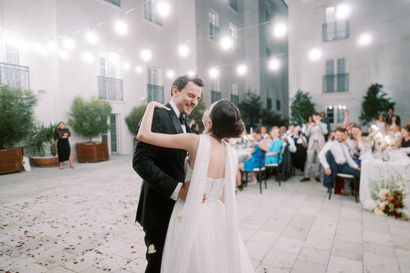 A bride and groom share their first dance under string lights at an outdoor wedding reception, surrounded by seated guests and a festive atmosphere.