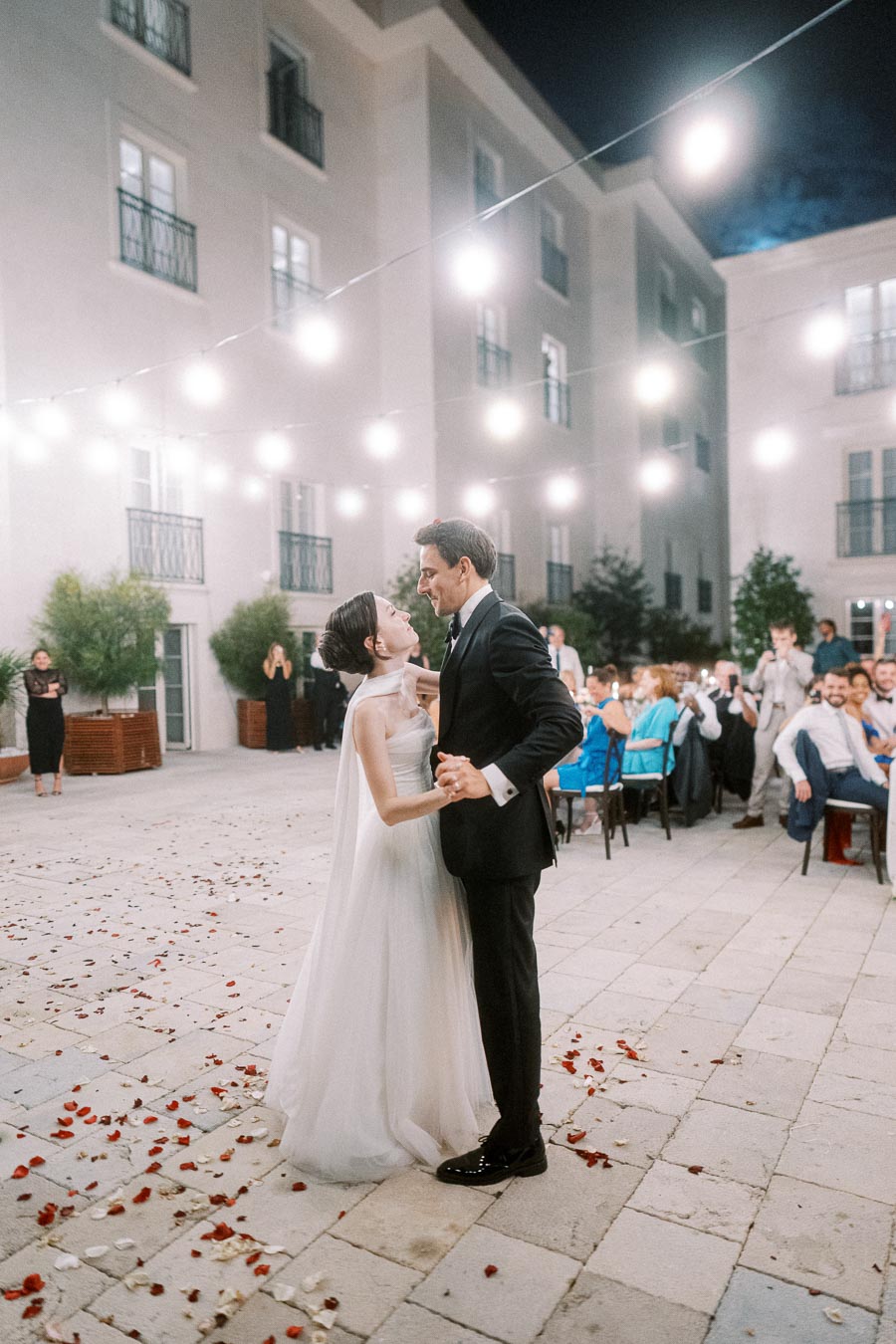 Elegant bride and groom share a romantic first dance under string lights at a nighttime outdoor wedding reception, with guests watching in the background.