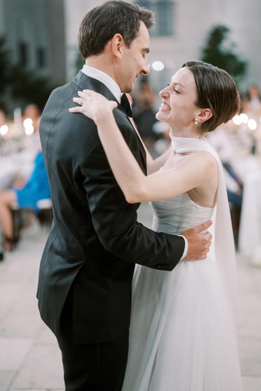 A bride and groom share a joyful first dance at their wedding reception, smiling and embracing on a softly lit venue.