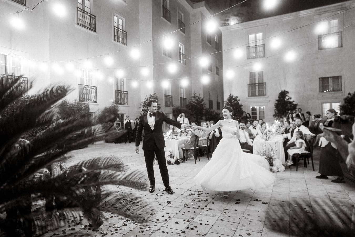 Black and white photo of a bride and groom dancing under string lights at an outdoor evening wedding reception, surrounded by seated guests and decorated tables.