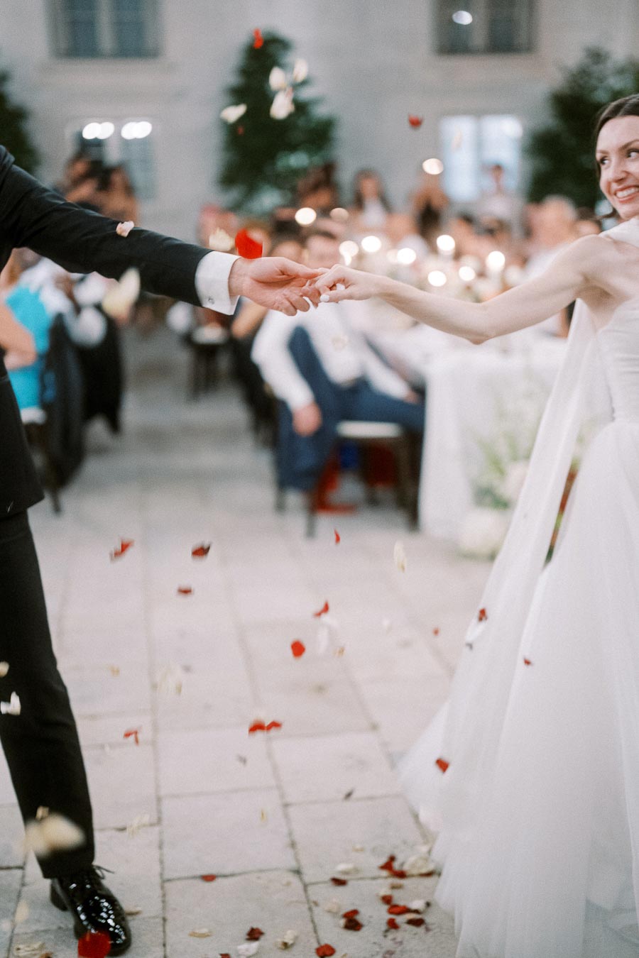 Wedding couple dancing hand in hand amidst falling rose petals, surrounded by guests, creating a romantic and joyful atmosphere at an outdoor reception.