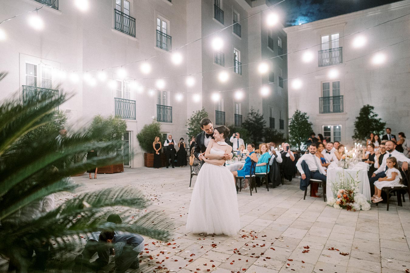 Outdoor wedding reception with bride and groom dancing under string lights, surrounded by seated guests and lush greenery.