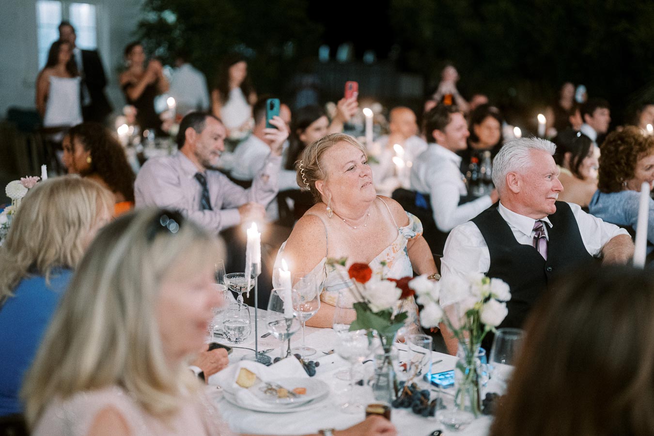 A lively group of people enjoying a candlelit dinner at an outdoor event, with elegantly set tables, floral arrangements, and a warm ambiance.