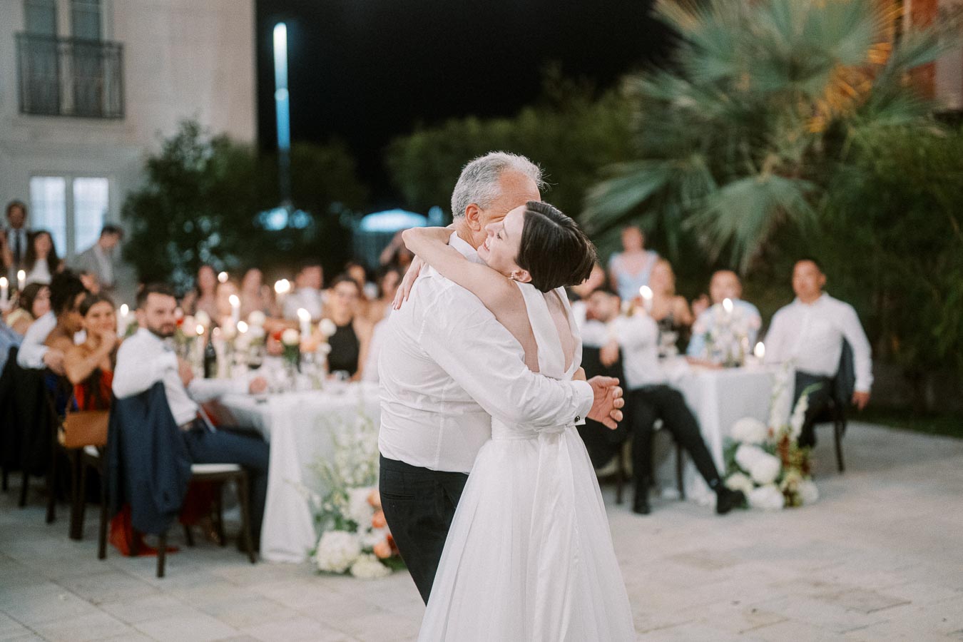 A bride and an older man embrace during a dance at an outdoor wedding reception at night, surrounded by seated guests and candlelit tables.
