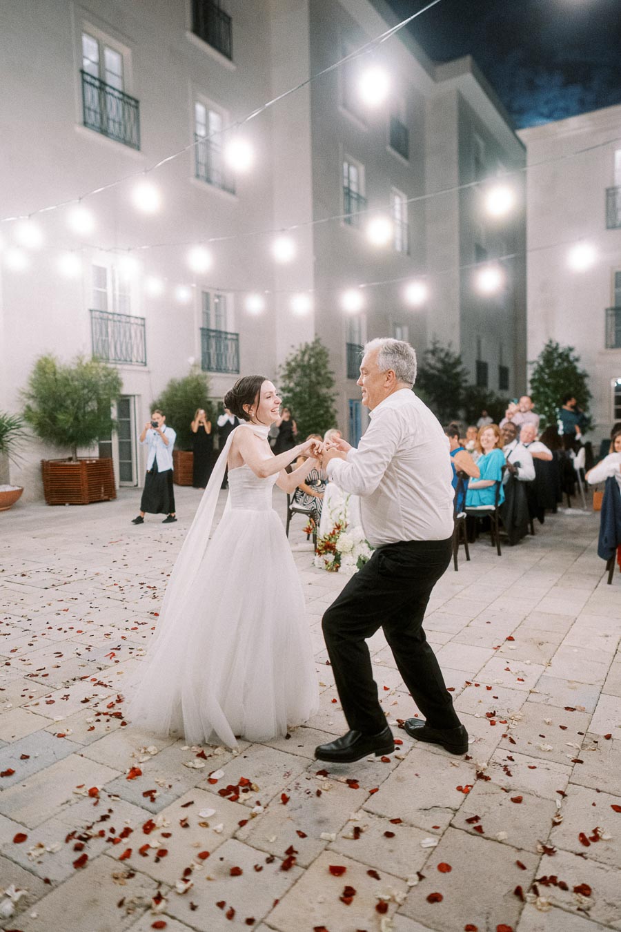 Father and bride dancing joyfully at an outdoor wedding reception adorned with string lights and rose petals, surrounded by seated guests.