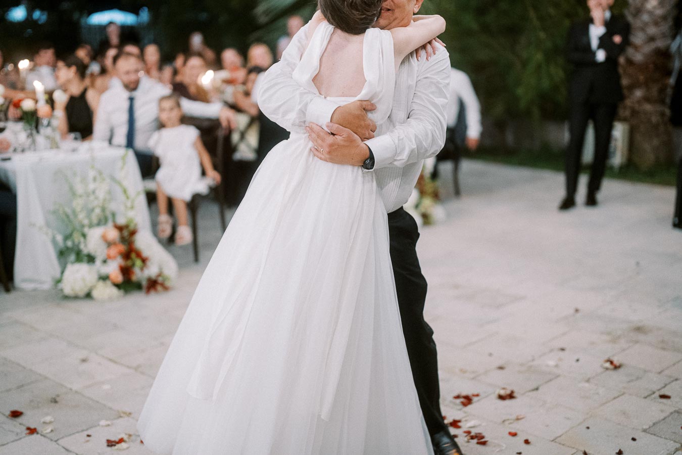 A bride in a white dress and a groom embrace during their first dance at an outdoor wedding reception, surrounded by guests seated at tables adorned with flowers.