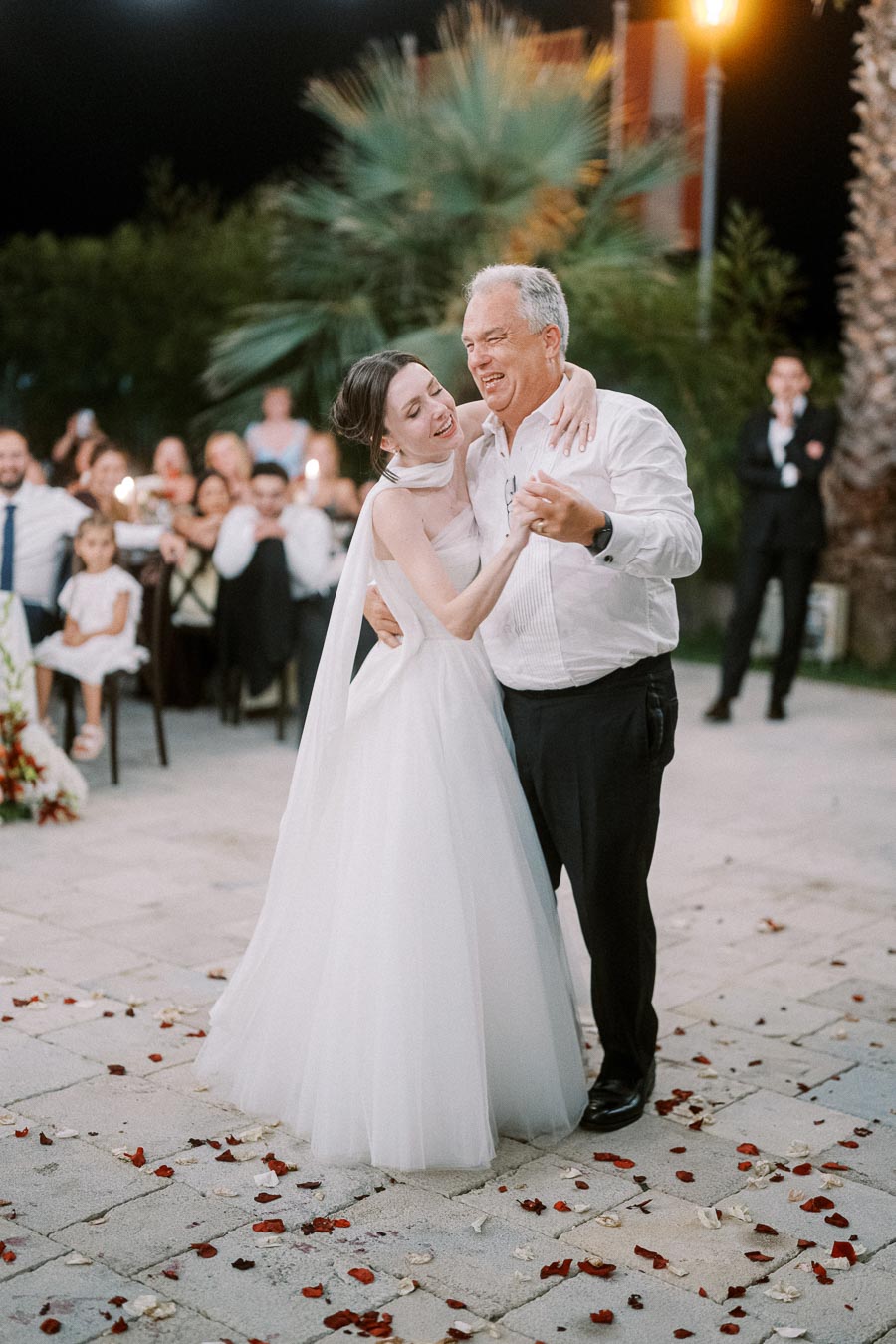 A bride in a white gown smiling while dancing with her father at an outdoor wedding reception, surrounded by guests and rose petals on the ground.