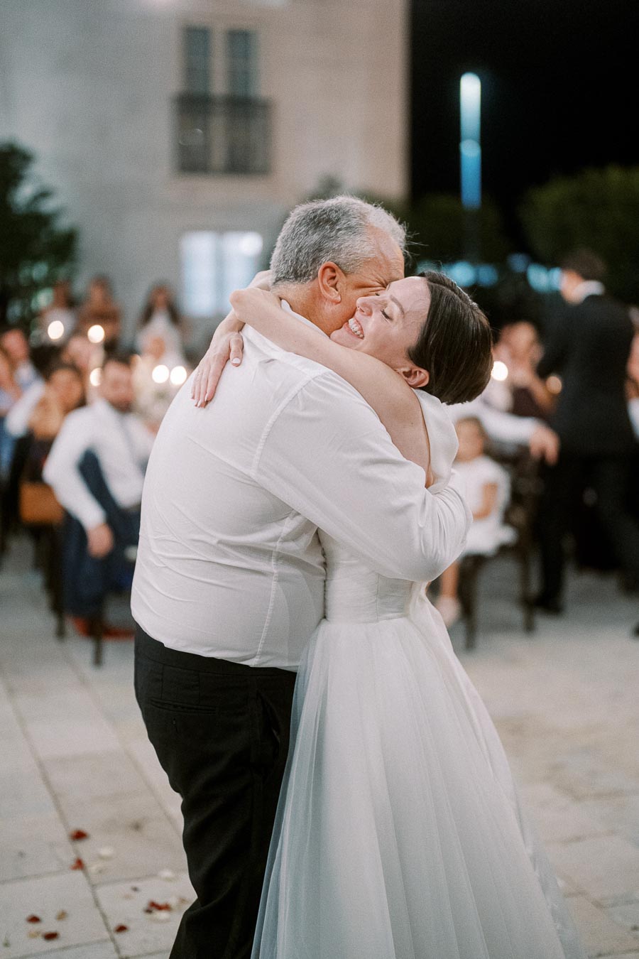 Father-daughter dance at a wedding reception, with the bride in a white dress smiling joyfully as she embraces her father in a warm hug under soft evening lighting.