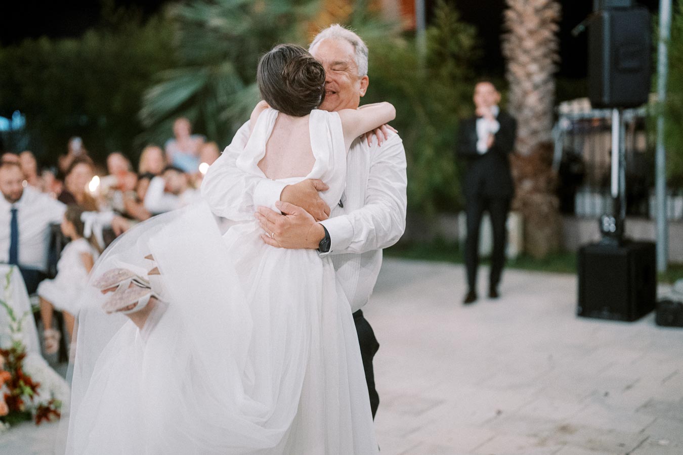 A joyous wedding moment as a bride in a white gown is lifted and embraced by an older man, surrounded by guests and greenery outdoors.