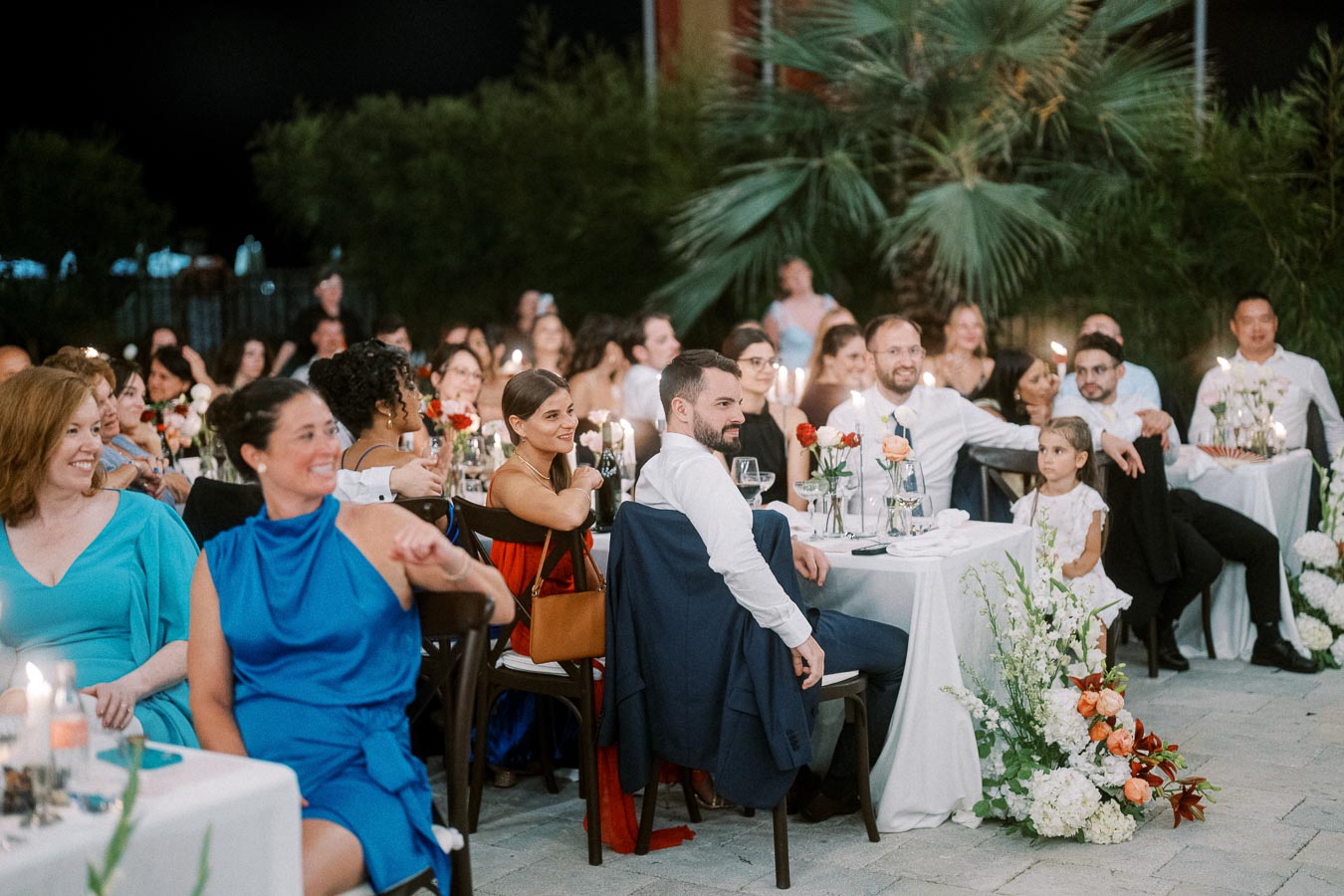 A diverse group of well-dressed guests enjoying an outdoor evening wedding reception, sitting at elegantly decorated tables with flowers, candles, and ambient lighting.