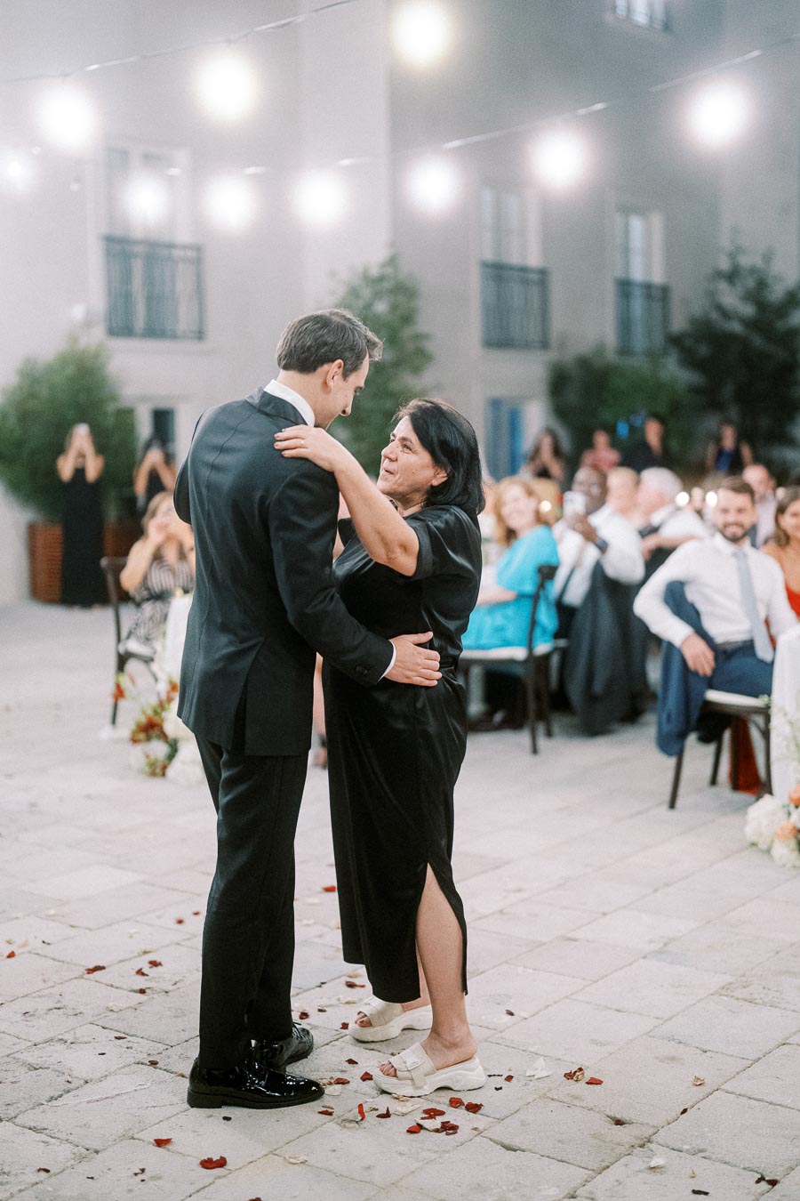A man in a suit dancing with a woman in a black dress at an outdoor wedding reception, with string lights and seated guests in the background.