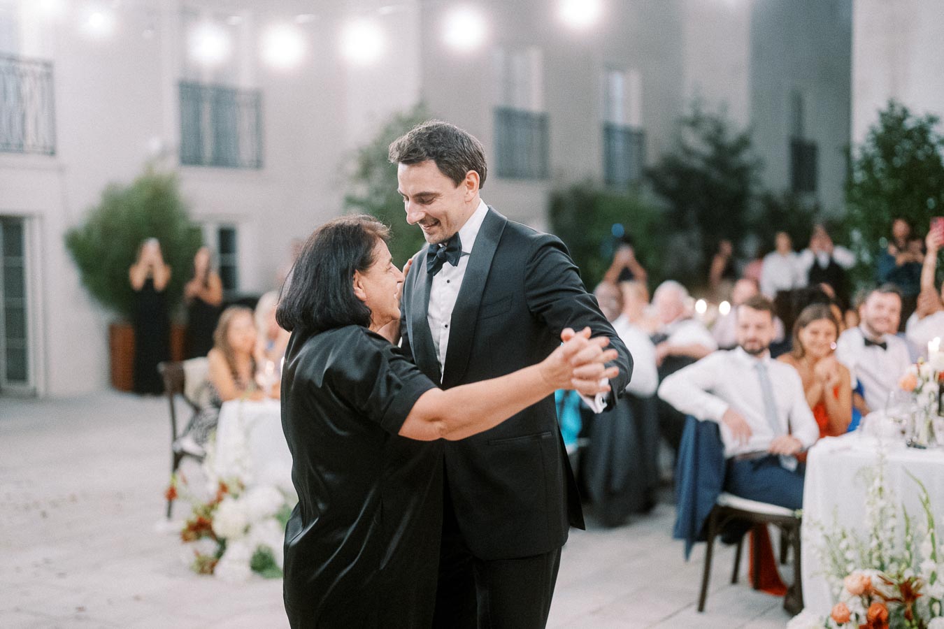 A groom in a black tuxedo dancing with a woman in an elegant black dress at an outdoor wedding reception, surrounded by seated guests and floral decorations.