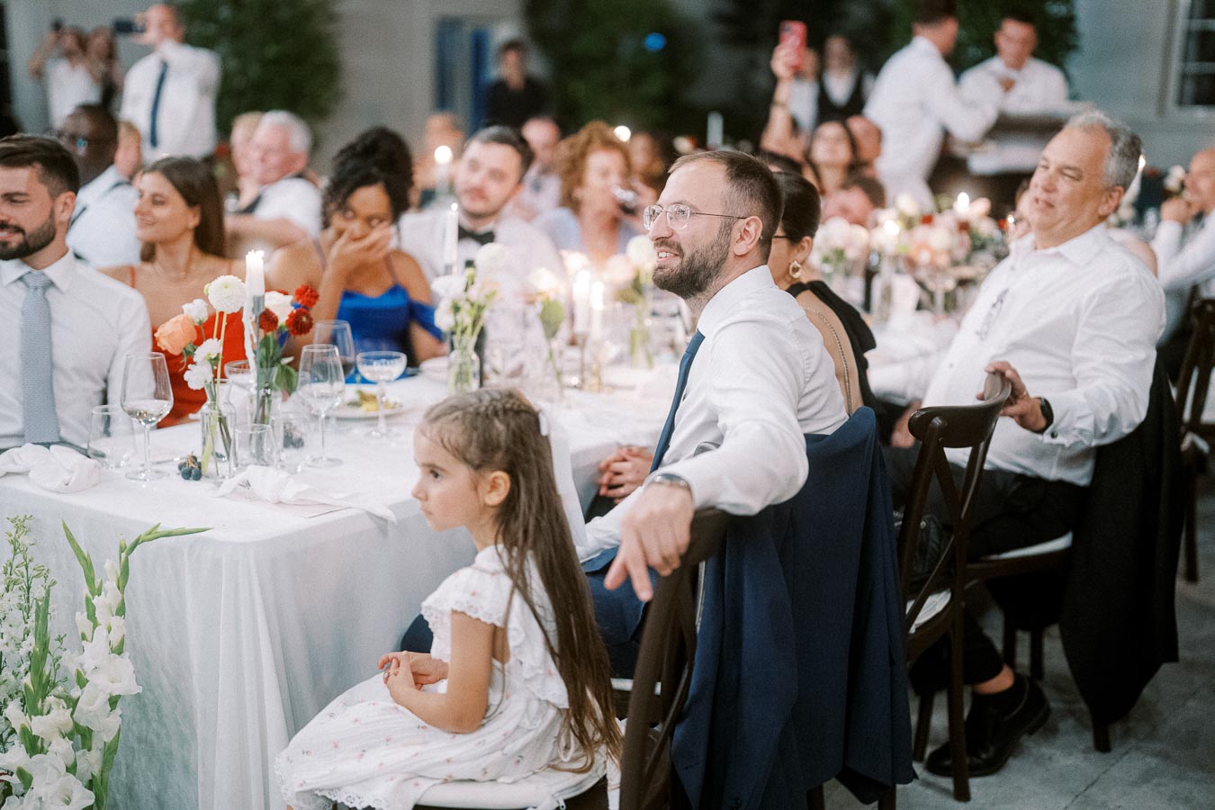 Guests enjoying a formal wedding reception, seated around a candlelit table with floral arrangements. A young girl in a white dress is seated at the front, surrounded by elegantly dressed attendees.