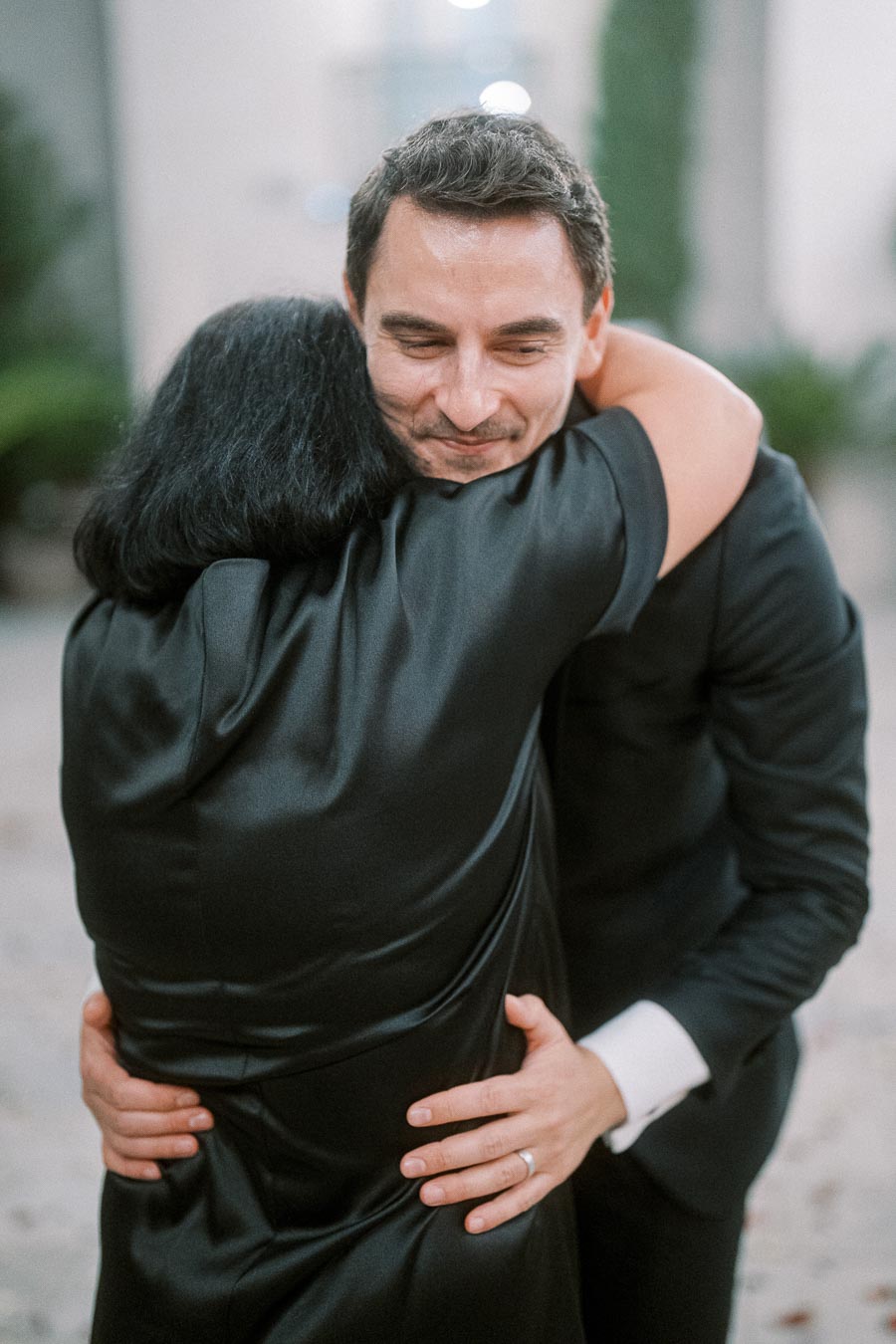 Man in a black suit warmly hugging someone, showing a joyful facial expression, in an outdoor setting with a blurred background.