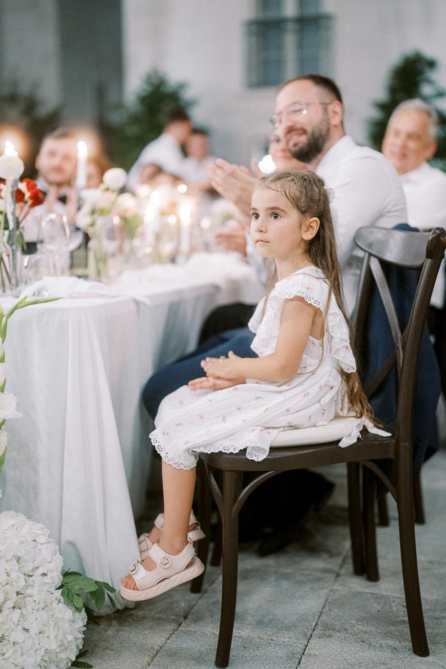 Young girl in white dress sitting at a dining table during an outdoor event, surrounded by guests and candlelight ambiance