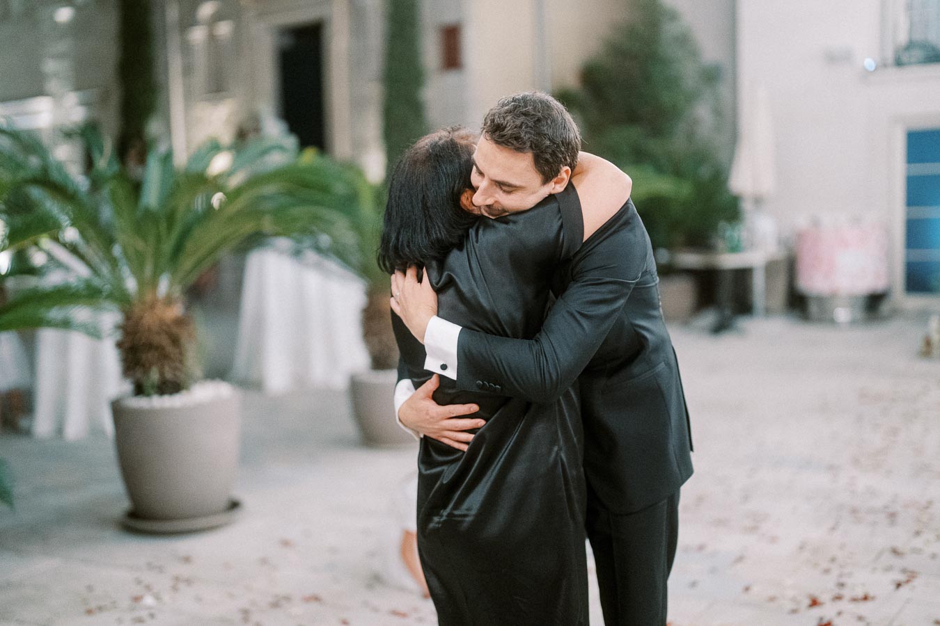 A man in a suit hugs a woman in a black dress warmly at an outdoor event, surrounded by greenery and potted plants.