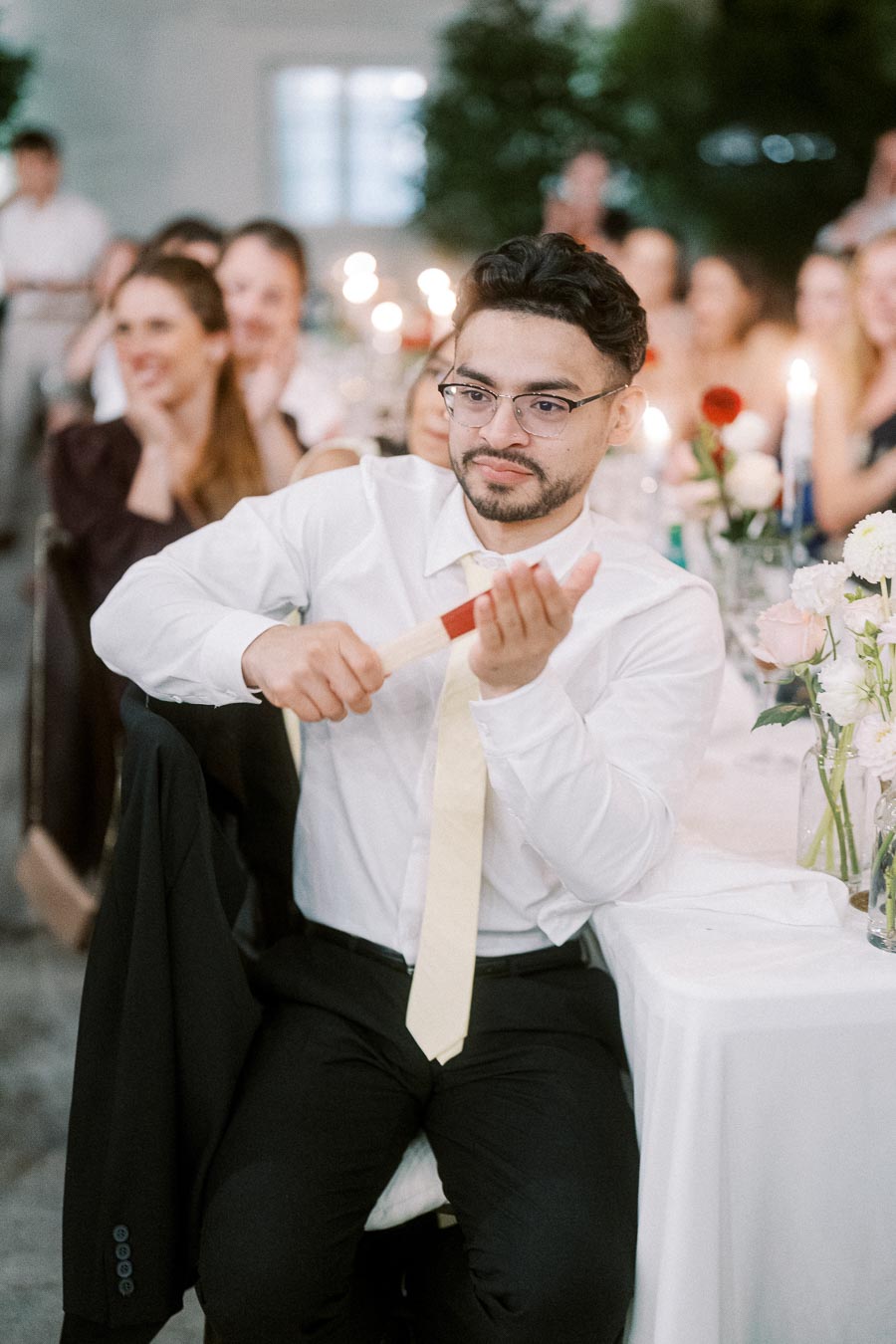 A person in formal attire smiling and interacting at a lively event, sitting at a table adorned with candles and flowers, surrounded by people in a celebratory atmosphere.