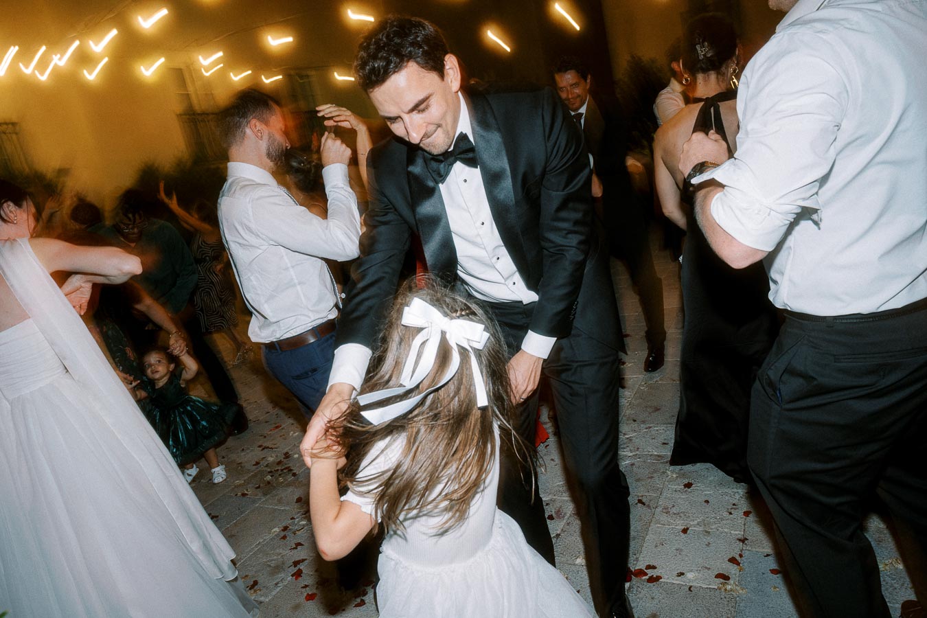 Wedding celebration with guests dancing energetically, featuring a man in a black tuxedo twirling a young girl in a white dress with a bow, surrounded by other elegantly dressed attendees under warm string lights.