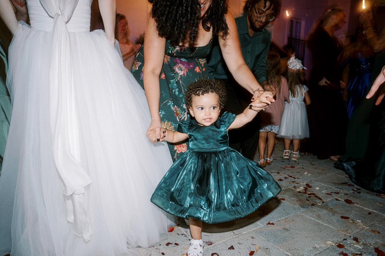 A young girl in a teal dress being assisted as she walks at a lively wedding reception, surrounded by guests in elegant attire.