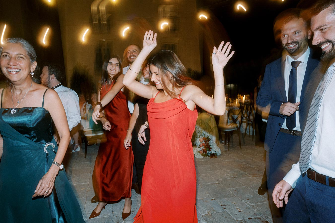 A group of people joyfully dancing at a lively evening event, with a woman in a red dress leading the celebration. Guests in formal attire smile and enjoy the outdoor party atmosphere under soft, ambient lighting.