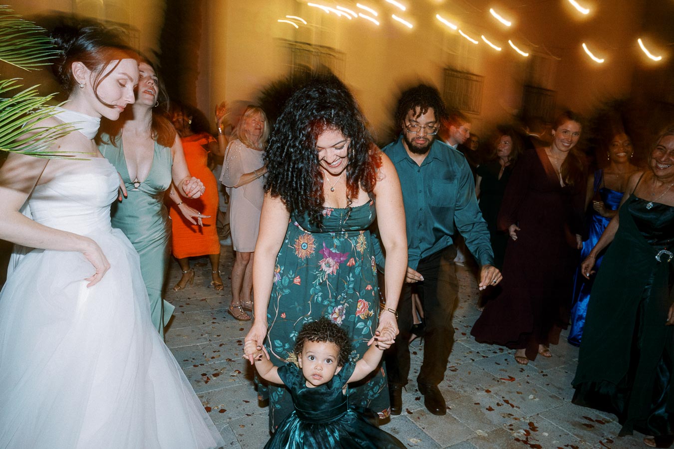A joyful wedding dance floor scene with guests in colorful attire, including a woman in a floral dress holding hands with a young child in a green dress, surrounded by smiling and dancing friends and family under soft ambient lighting.