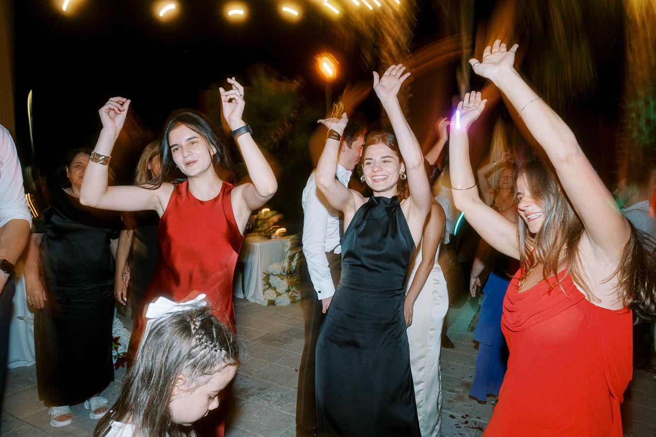 A group of people at a celebration dancing energetically under colorful lights, with women wearing elegant dresses enjoying the night.