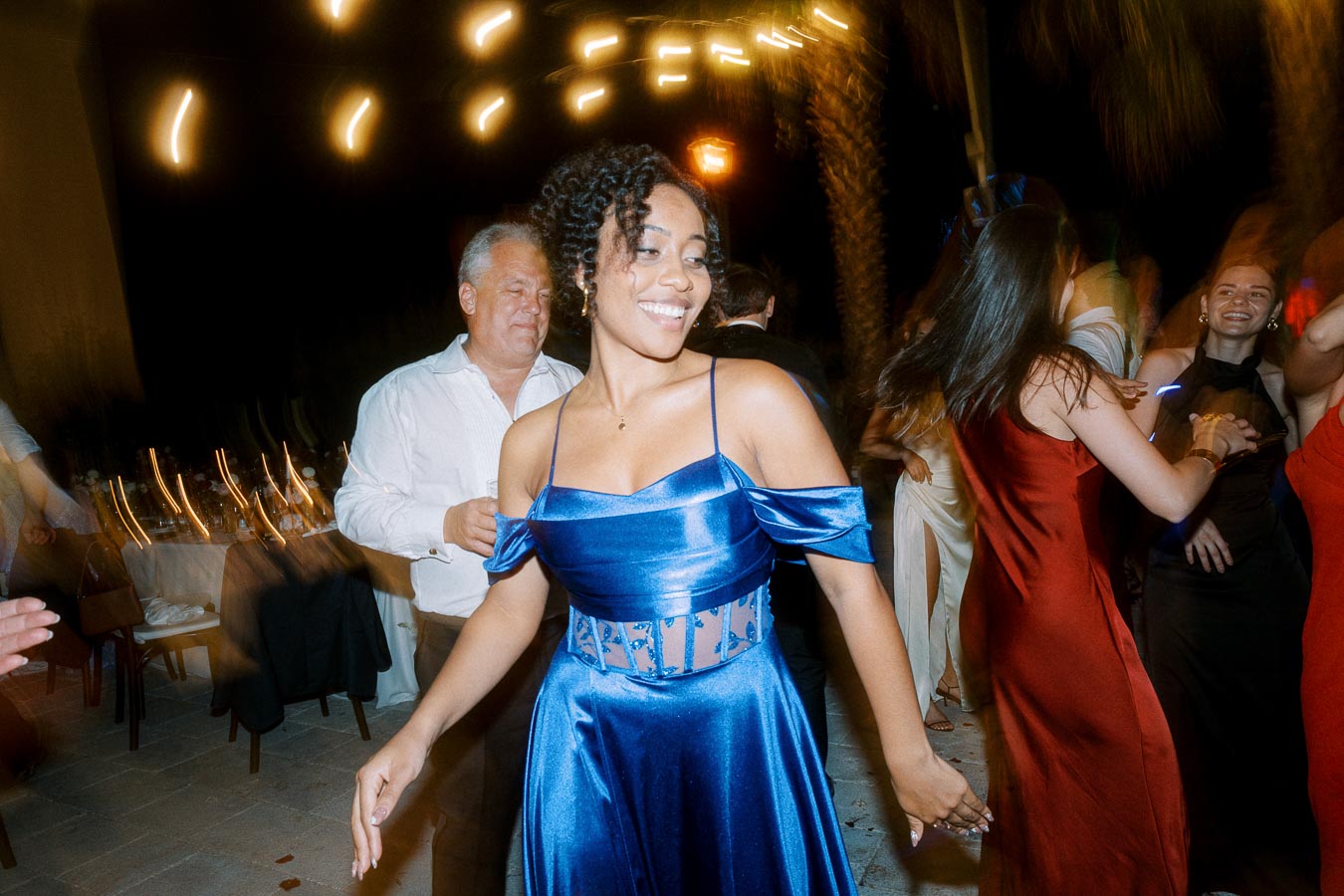Young woman in a blue dress dancing joyfully at a lively outdoor event, surrounded by people in elegant attire enjoying the celebration.