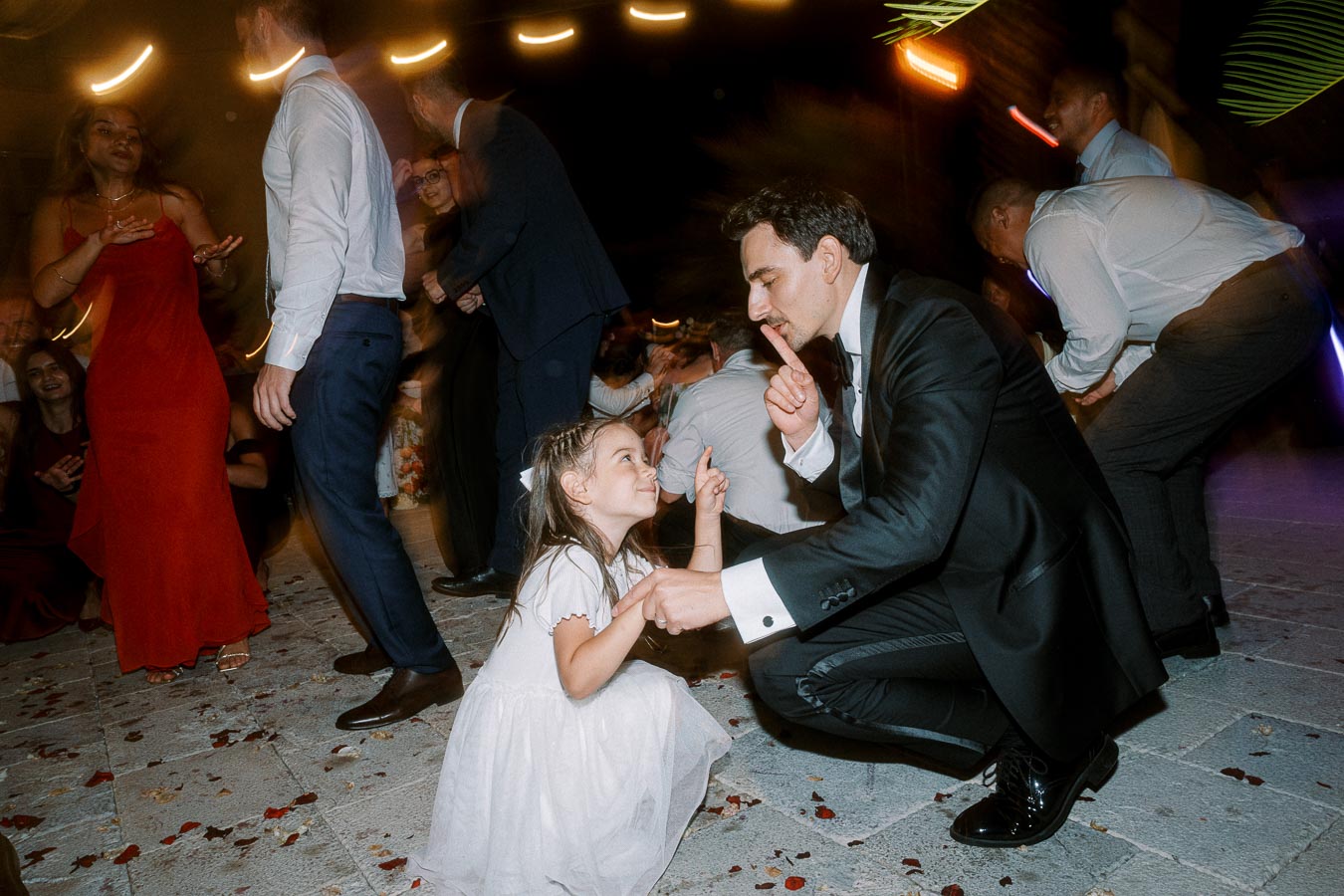 A man in a suit crouches and smiles at a young girl dressed in white while dancing at a lively wedding reception, surrounded by other guests.