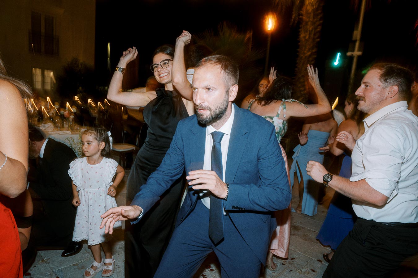 A lively wedding reception with guests dancing energetically under the night sky, featuring elegantly dressed individuals in suits and gowns, along with a young child in a white dress, capturing a joyous celebration atmosphere.