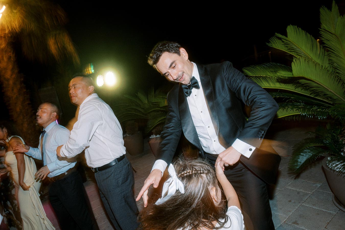 A man in a tuxedo dances joyfully with a child at an outdoor evening event, surrounded by other people dressed formally, with palm trees and bright lights in the background.