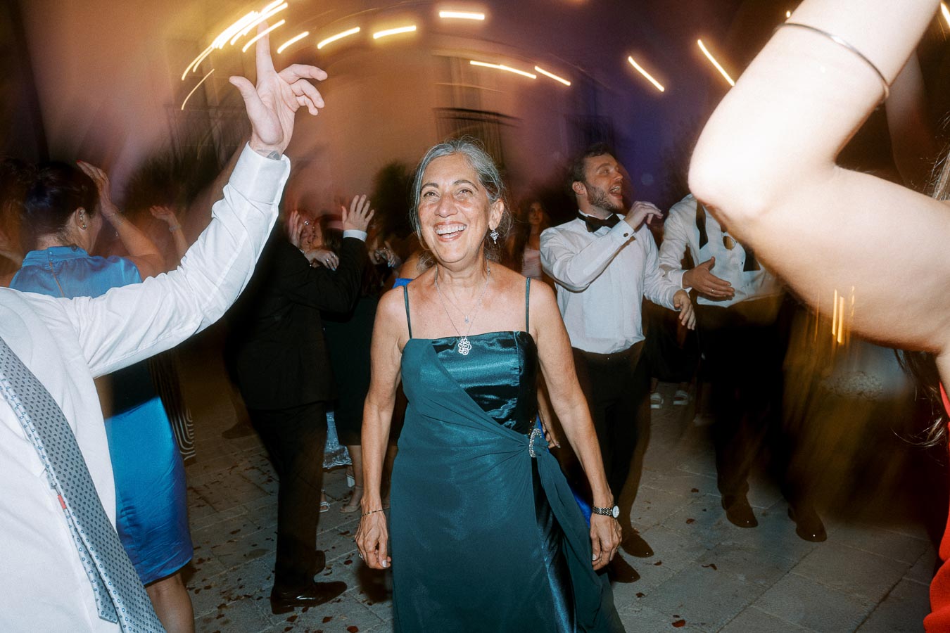 Elderly woman joyfully dancing at a lively outdoor celebration, surrounded by guests in formal attire with vibrant light trails in the background.