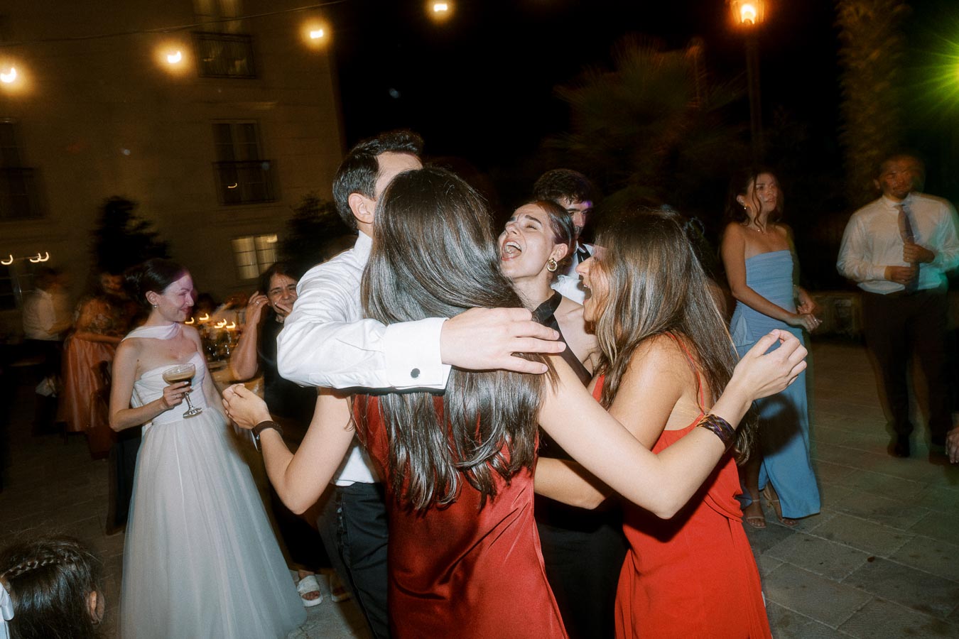 Group of friends joyfully dancing and celebrating at an outdoor evening event, dressed in formal attire with string lights in the background.