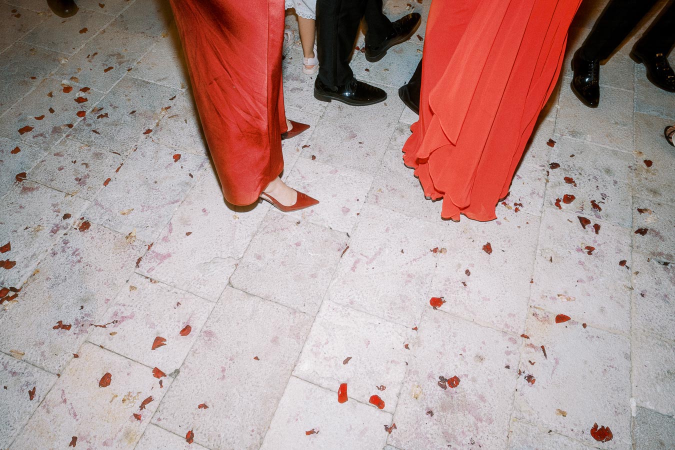 Guests in elegant red dresses and formal black shoes standing on a stone floor scattered with red rose petals.