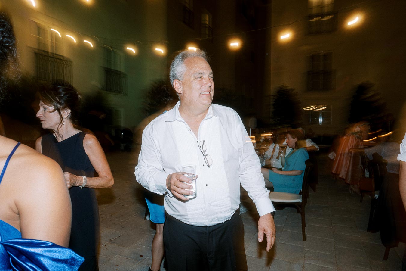 A middle-aged man wearing a white shirt holds a glass in an outdoor evening event surrounded by people and soft lights.
