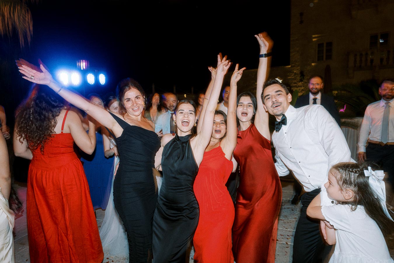 Group of people joyfully celebrating at an evening party, with women wearing elegant dresses and a man in a white shirt with a bow tie, surrounded by colorful lights and a festive atmosphere.