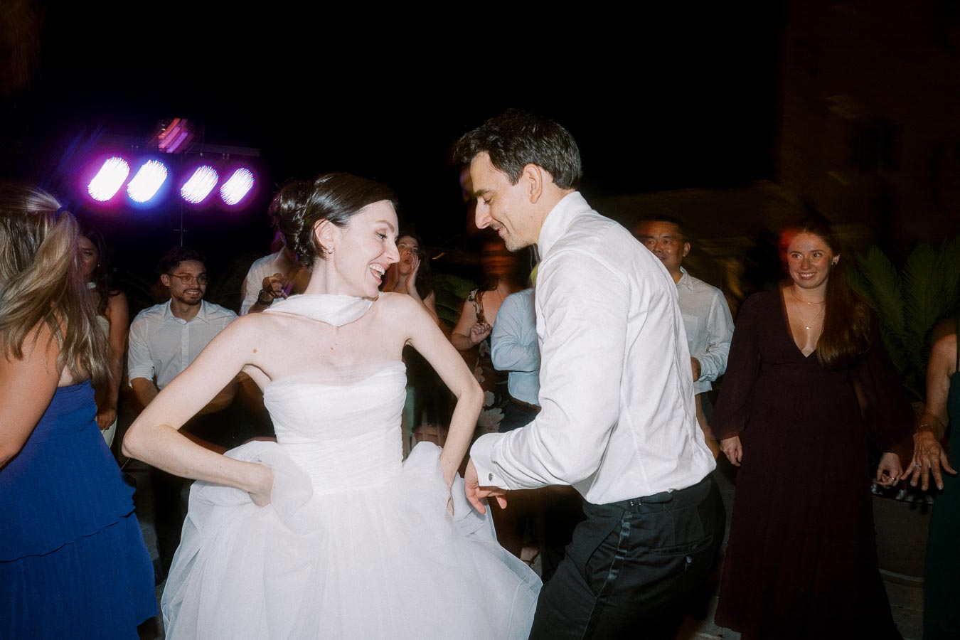 Newlywed couple dancing joyfully at their wedding reception, surrounded by friends and colorful lights.