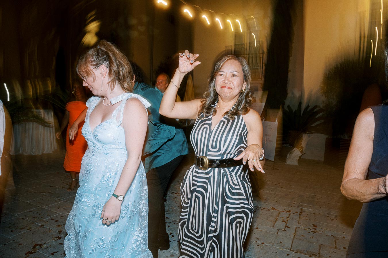A lively scene at a nighttime outdoor event, featuring several people dancing joyfully. The woman in the foreground wears a striped dress, raising her hand in a cheerful gesture, while another woman in a blue gown dances nearby. The ambient lighting and blurred motion capture the vibrant atmosphere.