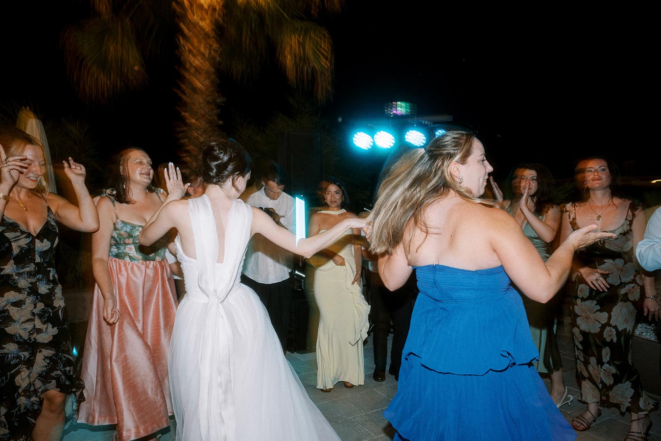 Group of women dancing joyfully at an outdoor evening event, wearing colorful dresses, with festive lights in the background.