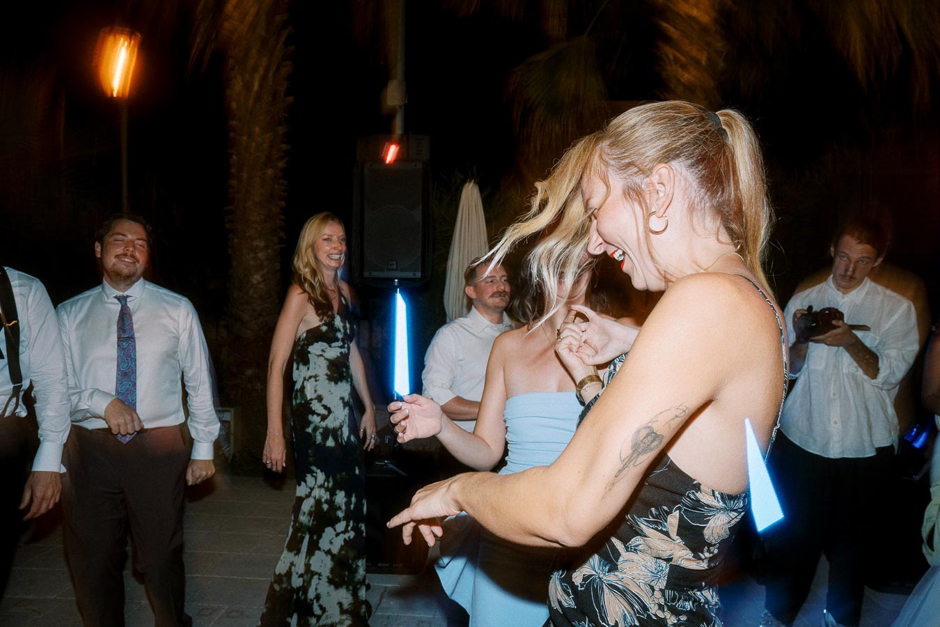 A group of people enjoying a lively dance party at night, with a woman in a patterned dress smiling and moving energetically, surrounded by others in formal attire and vibrant lighting.