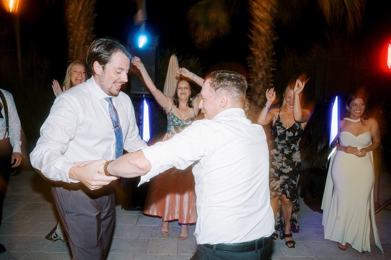 A group of people joyfully dancing at an outdoor night event, featuring well-dressed individuals in formal attire, with vibrant lights and palm trees in the background.