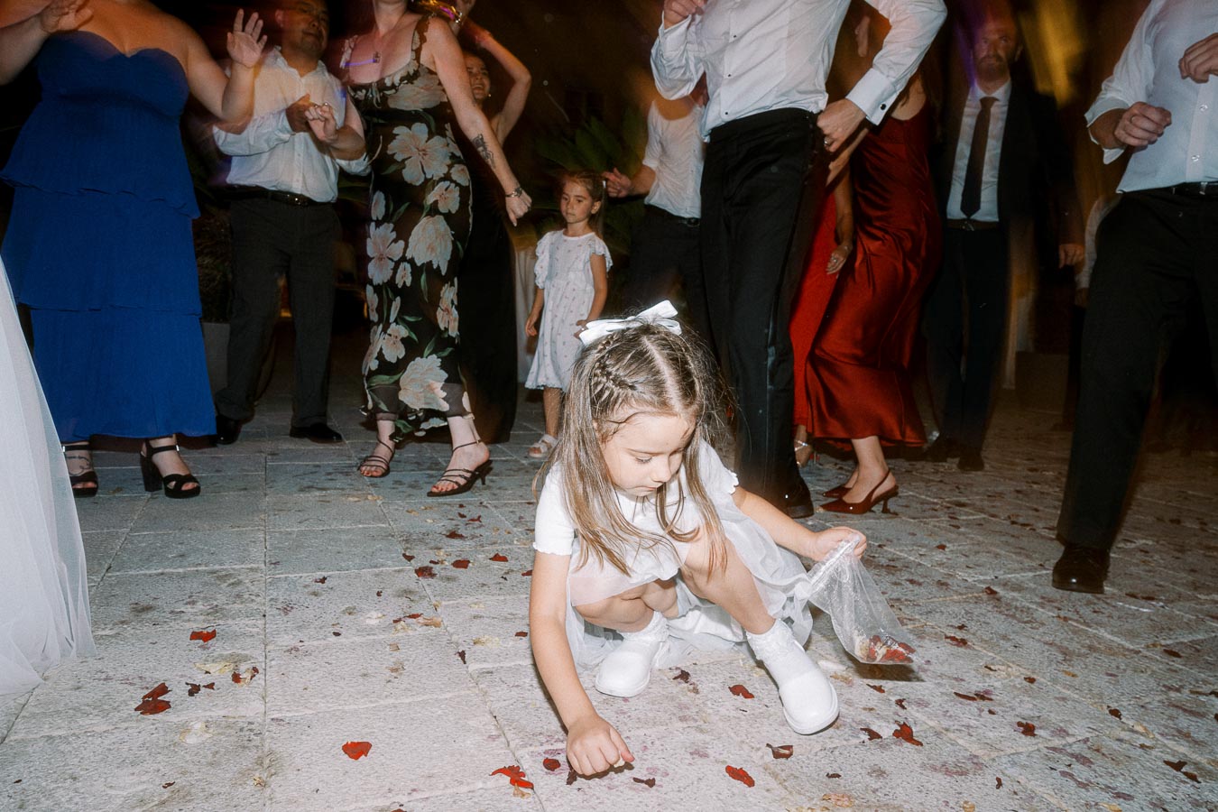 A young girl in a white dress picks up petals on a dance floor during a lively wedding celebration, surrounded by dancing adults in colorful attire.