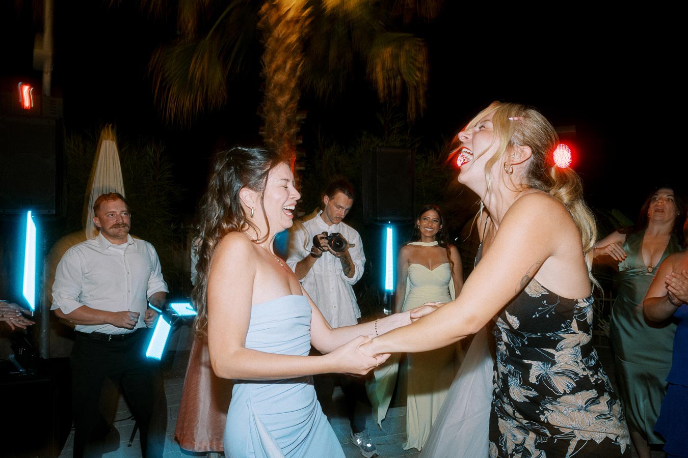 A joyful wedding celebration with two women laughing and dancing together under evening lights, surrounded by smiling guests in elegant attire. A photographer is capturing the moment, creating a lively and festive atmosphere.