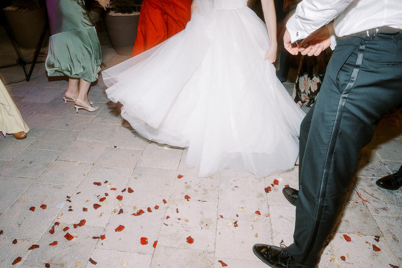 Guests dancing at a wedding reception, featuring a bride in a flowing white gown and other attendees in colorful attire, with scattered red rose petals on the tile floor.