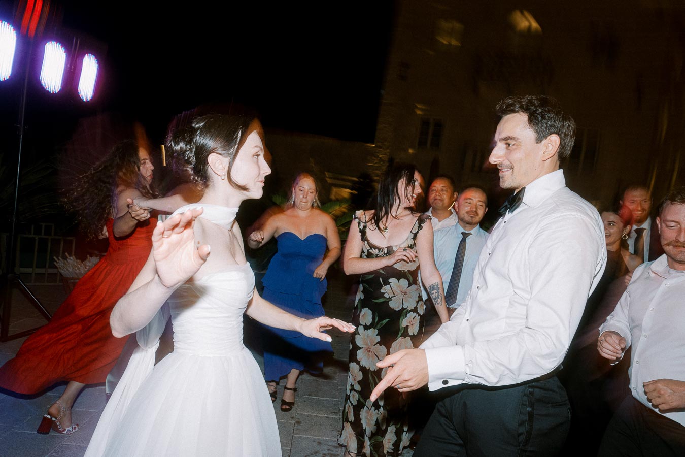 Elegant bride and groom dancing joyfully with guests at a lively night wedding reception, surrounded by friends in colorful evening attire.
