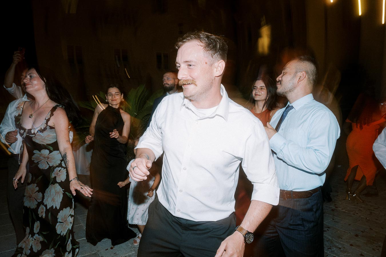 A group of people enjoying a lively dance at a nighttime event, with one person in a white shirt leading the moment, surrounded by others dressed in formal and semi-formal attire, set against a dimly lit background.