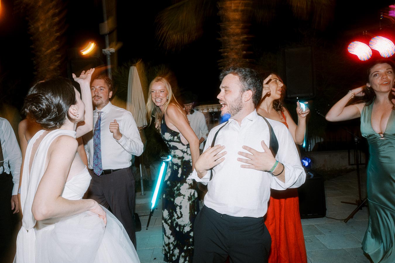 A group of people energetically dancing and enjoying a nighttime wedding reception, dressed in formal attire, with colorful lights in the background.