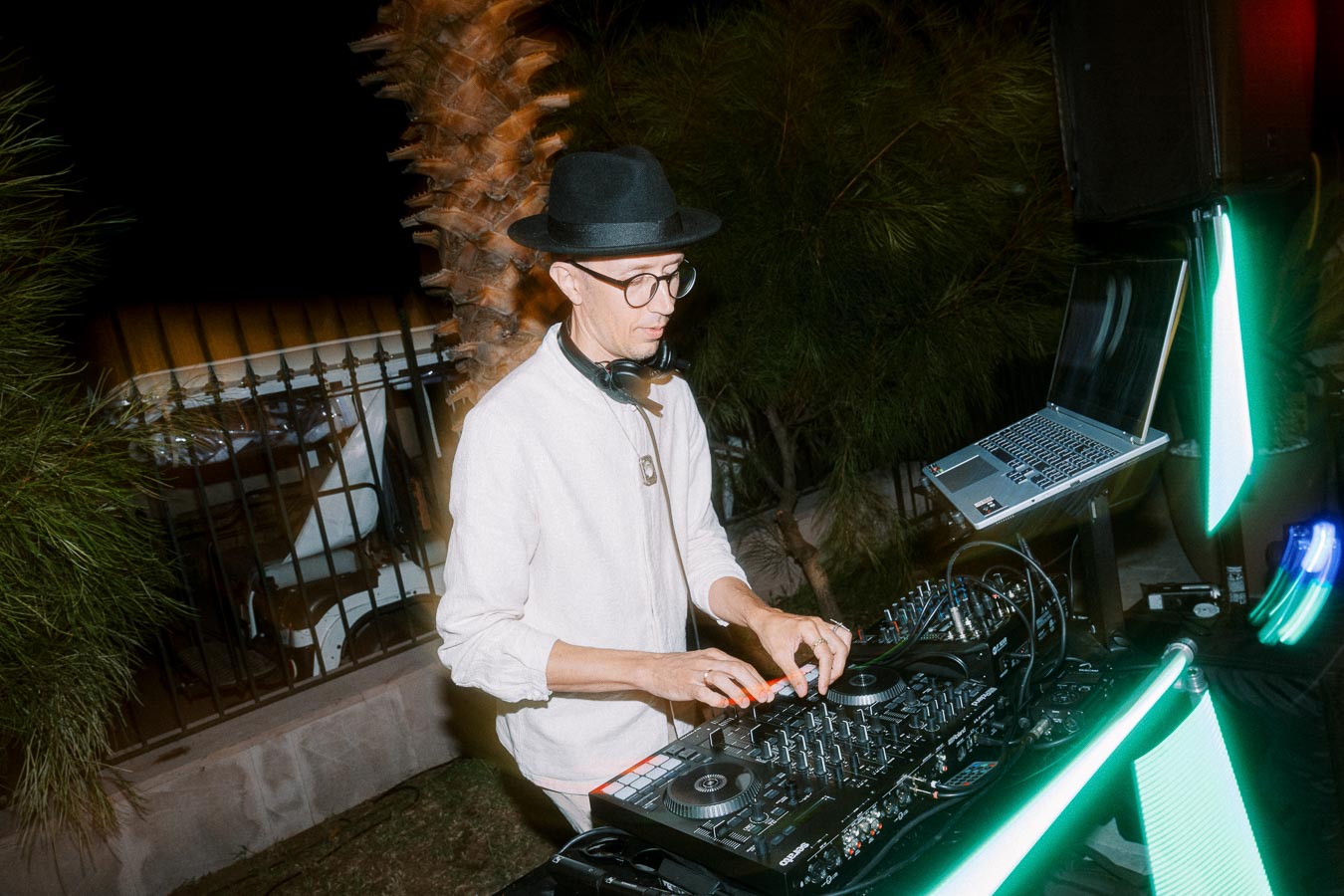 A DJ wearing a hat and glasses mixes music on a turntable setup outdoors at night, surrounded by greenery and LED lights.