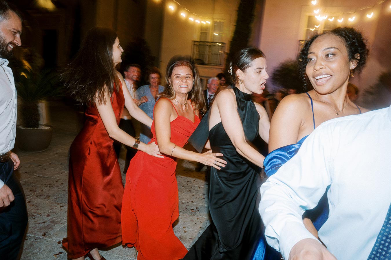 Group of people joyfully dancing in a conga line at a lively evening party, with festive lights illuminating the outdoor venue and participants dressed in formal attire, highlighting a celebratory atmosphere.