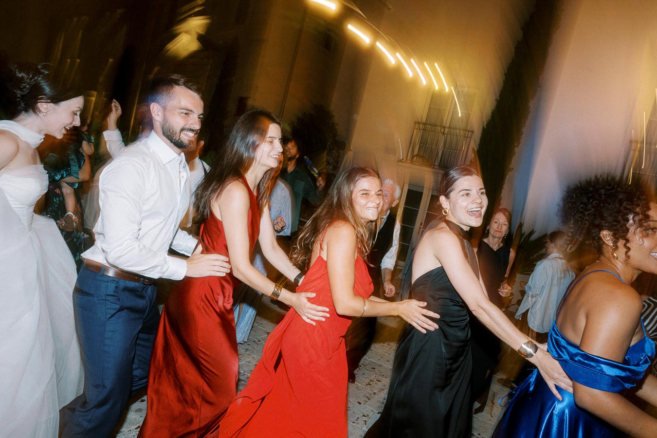 A group of joyful people in formal attire dancing at a vibrant party, with women in elegant dresses and a man in a white shirt, all smiling and enjoying the festive atmosphere.