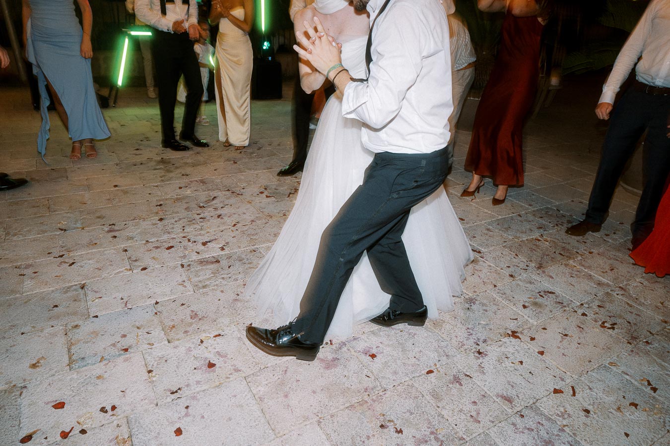 A bride and groom dancing energetically on a stone patio during a wedding reception, surrounded by elegantly dressed guests.